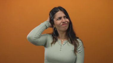 Kadın eli başının üzerinde ifade düşünüyor, turuncu arka planda rahat giyinmiş, kafa karışıklığı ve tefekküre dalmış.