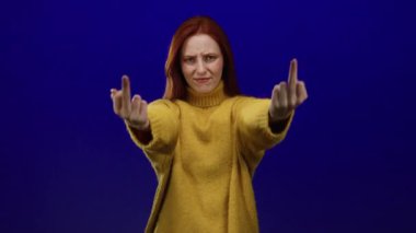 Sarı kazaklı bir kadın, mavi bir arka plana karşı saldırgan bir el hareketi sergiliyor. Küstah bir ifadeyle, kırmızı saçlar, canlı mavi duvara karşı duruyor. Asi havasını arttırıyor..