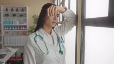 Doktor kadın, bir kliniğin penceresinin yanında steteskopla düşünceli görünüyor. Hastane ortamını inceliyor gibi görünüyor..