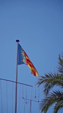 Valencia bayrağı açık mavi gökyüzüne karşı palmiye ağacıyla dalgalanıyor, İspanyol bölgesinin güneşli açık hava güzelliğini ve canlı kültürünü simgeliyor..