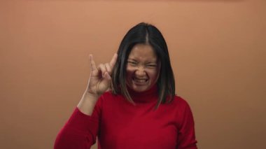 Kırmızı kazaklı kadın kahverengi duvara karşı kaya hareketi yapıyor, neşe ve enerji ifade ediyor, canlı bir ifade ve gençlik coşkusu sergiliyor..