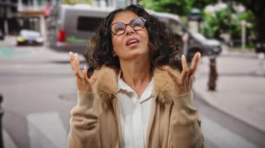 Kıvırcık saçlı bir kadın şehir sokaklarında gözlük ve açık havada kalın bir palto giyer..