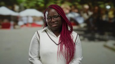 Pembe örgülü ve gözlüklü Afro-Amerikan kadın şehir manzarasında bulanık kalabalıkla düşünceli bir ifade sergiliyor..