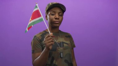 Kamuflaj şapkası ve künyesi takan kadın asker, stüdyoda mor arkaplan ve gurur görevine sahip surinam bayrağı taşıyor..