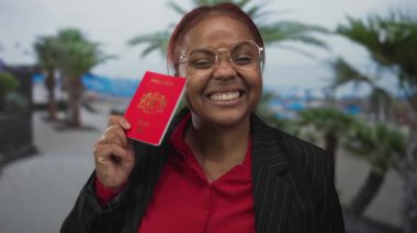 Yuvarlak gözlüklü kadın sırıtıyor ve plajda palmiye ağaçlarıyla kaplı kırmızı Malezya pasaportunu tutuyor; seyahat keyfi kutlaması.