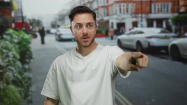 Genç sakallı bir adam heyecanlı bir şehir caddesinde arabaları ve bulutlu gökyüzünün altından geçen insanları işaret ediyor..