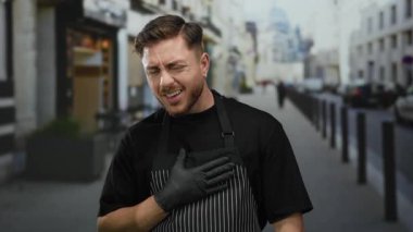 Önlüklü genç adam şehir sokağında dikiliyor, düşünceli bir ifadeyle eldiven takıyor, şehir hayatını yakalıyor..