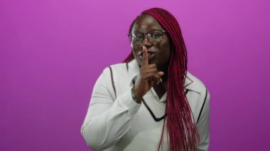Gözlük ve beyaz süveter giyen, soyutlanmış pembe bir arka plana karşı sessiz kalmak için el kol hareketi yapan Afrikalı Amerikalı bir kadın..
