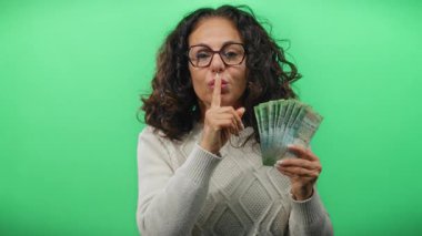 Elinde Rus rublesi taşıyan orta yaşlı bir kadın canlı yeşil bir arka plana karşı sessizlik işareti veriyor. Gizemli bir finans sahnesi yaratıyor..