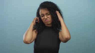 Bukleli ve gözlüklü bir kadın mavi arka planda duruyor, endişeli bir ifadeyle kulaklarına dokunuyor, siyah bluz giyiyor, izole edilmiş ve hoşnutsuz görünüyor..