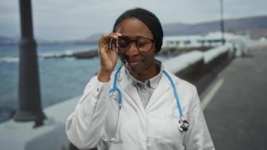 Steteskoplu beyaz önlüklü bir kadın, bulanık bir okyanus geçmişine karşı deniz kenarında neşeli bir profesyonel izlenim sunuyor..
