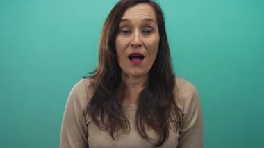 Canlı yeşil arka planda gülümseyen bir kadın uzun siyah saçlı ve bej renkli kazaklı neşeli ve şaşırtıcı ifadeler sergiliyor..