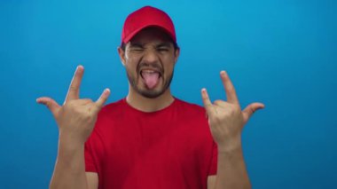 Stüdyoda parlak kırmızı şapkalı ve tişörtlü genç adam rock boynuzlarını diliyle gösteriyor..