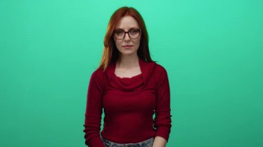 Kırmızı saçlı ve gözlüklü bir kadın yeşil duvara karşı kırmızı kazak giyiyor. Düşünceli ve kendinden emin ifade ediyor. Münferit bir ortam modern ve şık görünümü vurguluyor.