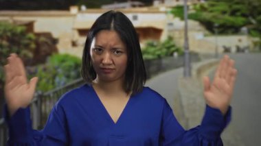 Genç bir kadın, parlak bir günde şehir caddesinde mavi bir tişört giyerek binaların ve yeşilliklerin olduğu duyguları yansıtan bir jest yapıyor..