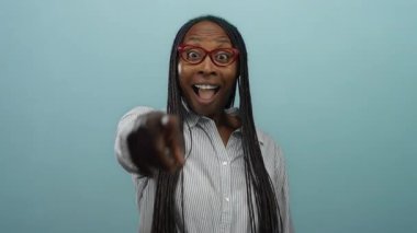 Kıvırcık saçlı, gülümseyen bir kadın mavi bir duvar arkasına işaret ederken neşeli bir ifade ve neşeli bir ruh hali sergiliyor..