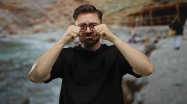 Deniz kenarında sakallı ve gözlüklü genç adam yaz aylarında bulanık arkaplanlı sahilde güneşli gökyüzünün altında üzgün görünüyor..