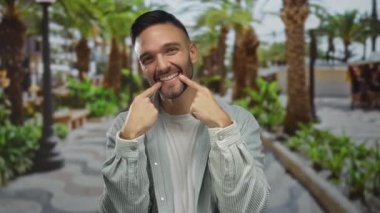 Palmiye ağaçlarıyla dolu yemyeşil bir park yolunda dikilirken gülümsemesini gösteren parmaklarıyla gülümseyen İspanyol genç adam.