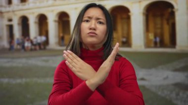 Genç Çinli bir kadın, üniversite ortamında çapraz kollarıyla x hareketi yapıyor, kırmızı bir kazak giyiyor, eski mimari geçmişinden duyduğu memnuniyetsizliği dile getiriyor..
