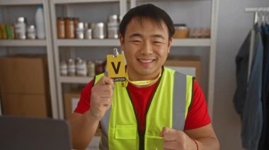 Gönüllü bir rozet ve güvenlik yeleği takan, bağış rafları, toplum ruhunu ve takım çalışmasını sergileyen, gülümseyen ve onaylayan bir hayır kurumundaki genç adam..