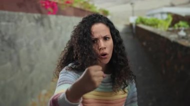 Açık havada, kıvırcık saçlı ve gökkuşağı süveterli bir kadın, yumruğuyla taştan duvar zeminine karşı öfkesini ve kararlılığını canlı kent ortamında çiçeklerle gösteriyor..