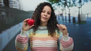 Açık havada elinde kırmızı bir kalple gülümseyen bir kadın şehir caddesinde renkli bir süveter giyerek, canlı bir şehir ortamında, pozitifliği ve aşkı öneriyor..