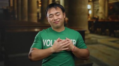 Gönüllü olarak etiketlenmiş yeşil gömlekli genç adam kilisede mutlu duruyor, ellerini göğsüne koyuyor, dini bir ortamda minnettarlık ve memnuniyeti ima ediyor..