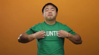 Yeşil gönüllü gömlekli bir adam turuncu bir duvarın önünde duruyor ve elleri göğsünde kendinden emin bir ifade sergiliyor..