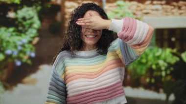 Açık havada neşeli bir şekilde gülümseyen bir kadın canlı sokak arka planında renkli bir süveter giyerek yemyeşil bir ortam yaratıyor.