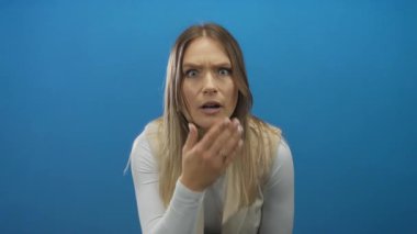 Ağzı mavi arka planda kaplayan kadın, geniş gözlü ve el hareketleriyle şok gösteriyor, beyaz bluz giyiyor, izole edilmiş portre tarzı..