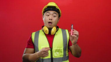 Güvenlik teçhizatlı genç adam kırmızı arka plana karşı vape cihazını işaret ederek güvenli bir şekilde neşeli bir ifadeyle ürünü gösteriyor.