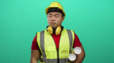 Asyalı genç bir adam inşaat malzemesi giyiyor yeşil arka planla ilgili planlar tutuyor mesleğini gösteriyor ve açık havada mimarlık işine odaklanıyor.