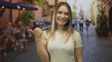 Şehir terasında gülümseyen bir kadın, arka planda insanlar ve gelişigüzel bir şehir kafesi ile baş parmağını yana uzatıyor..