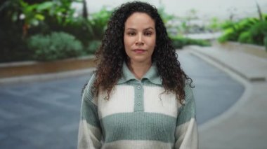 Kıvırcık saçlı genç bir İspanyol kadın sokakta kameraya bakıyor. Etrafı yemyeşil, neşeli bir ifadeyle çevrili. Neşeli, samimi bir anın altını çiziyor..