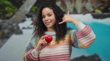 Bir otelde havuz kenarında kırmızı kalbi tutan, gülümseyen ve rahat bir şekilde güneşli gökyüzünün altında, açık havada renkli bir kazak giyen bir kadın..