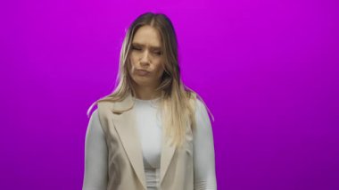 Beyaz ve bej renkli sarı saçlı kadın canlı mor bir arka plan duvarına karşı düşünceli duruyor. Düşünce ve belirsizlik ifadeleri sergiliyor..