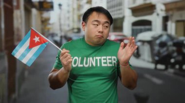 Elinde Küba bayrağı tutan genç bir adam. Şehir caddesinde yüzü belirsiz. Gönüllü tişörtü giyiyor.