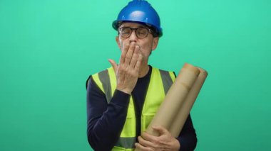 Mavi miğfer ve güvenlik yeleği giyen adam yeşil arka plana karşı yuvarlanmış planlar tutuyor..