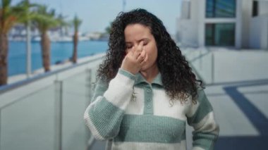 Plaj gezisindeki kadın sıcak süveteri ve okyanus altyapısı ile rahatsızlığını dile getiriyor. Güneşli bir günde palmiyelerin ve modern mimarinin huzurlu manzarasını yakalıyor..