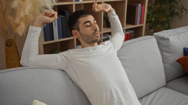 Genç İspanyol bir adam, rahat bir oturma odasında rahat bir kanepeye uzanıyor, etrafı kitaplar ve bitkilerle çevrili, evinde, davetkar bir apartman dairesinde rahatlama gösterisi yapıyor..