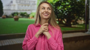 Dışarıda pembe tişörtlü bir kadın, arka planda yeşil bahçeli ve tarihi binalı bir sevinç ifadesi sergiliyor, mutluluğu, gençliği ve doğanın güzelliğini vurguluyor..