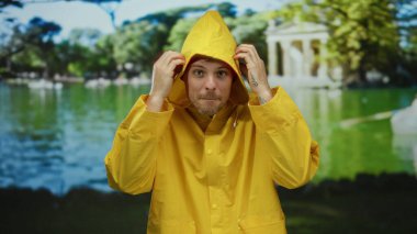Sarı yağmurluk giyen adam parlak bir günde yemyeşil ve sakin sularla dolu canlı bir parkta dalgın dalgın duruyor..