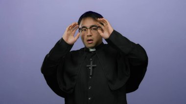 Papaz kıyafetli genç adam, izole bir mor arka planda, kaldırılmış eller ve kaldırılmış gözlüklerle, şok ve dini aktararak irkildi..