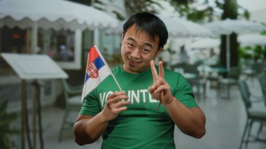 Terasta Sırp bayrağı taşıyan genç bir adam yeşil gönüllü tişörtü giyerken şehir ortamında neşeli bir ifade ve barış hareketi sergiliyor..