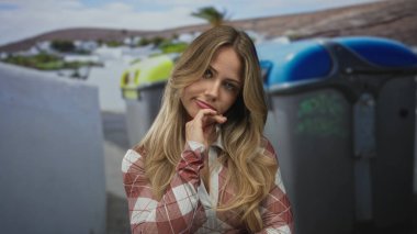 Çıplak kollu ve eli çenesinde olan kadın şehir caddesinde çöp bidonlarının yanında düşünürken, tefekkür merakı yansıtma havasında..