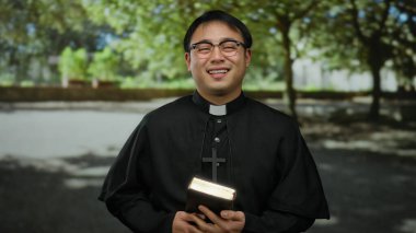 Elinde İncil 'le parkta gülümseyen Asyalı rahip, ağaçların gölgesinde siyah rahip kıyafeti giyiyor, neşe, maneviyat ve açık havada inanç taşıyor..