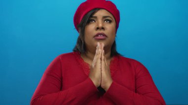 Kırmızı bereli ve kazaklı bir kadın, canlı mavi bir arka planda duruyor, umudunu ve inancını kapalı gözlerle ifade ediyor..