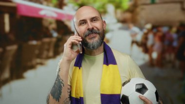 Futbol topu tutan ve şehir merkezinde telefonla konuşan yakışıklı kel adam sarı mor atkı takıyor spor tutkusu ve bağlantısını gösteriyor..