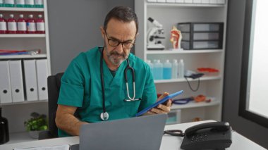 Modern klinikteki hasta kayıtlarını inceleyen erkek doktor tıbbi ekipmanları ve düzenli çalışma alanını gösteriyor..