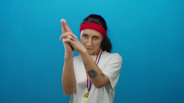 Madalyalı genç kadın, mavi arka planda kırmızı saç bandı takan, kendine güveni ve eğlencesi olan şakacı bir şekilde parmak tabancalarıyla poz veriyor..